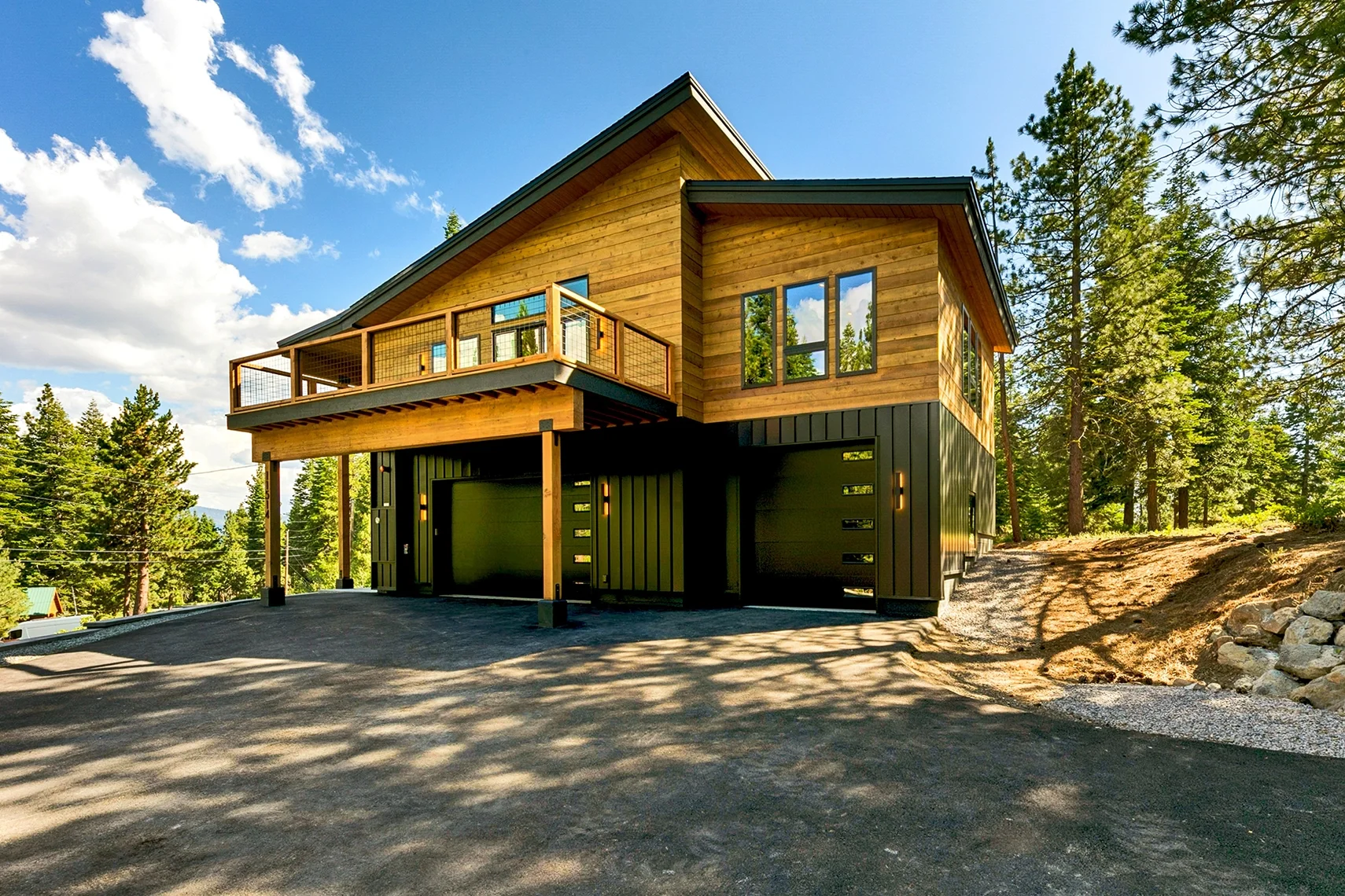 Modern wood-clad mountain home with a large second-story deck, black garage doors, and tall pine trees under a bright blue sky.