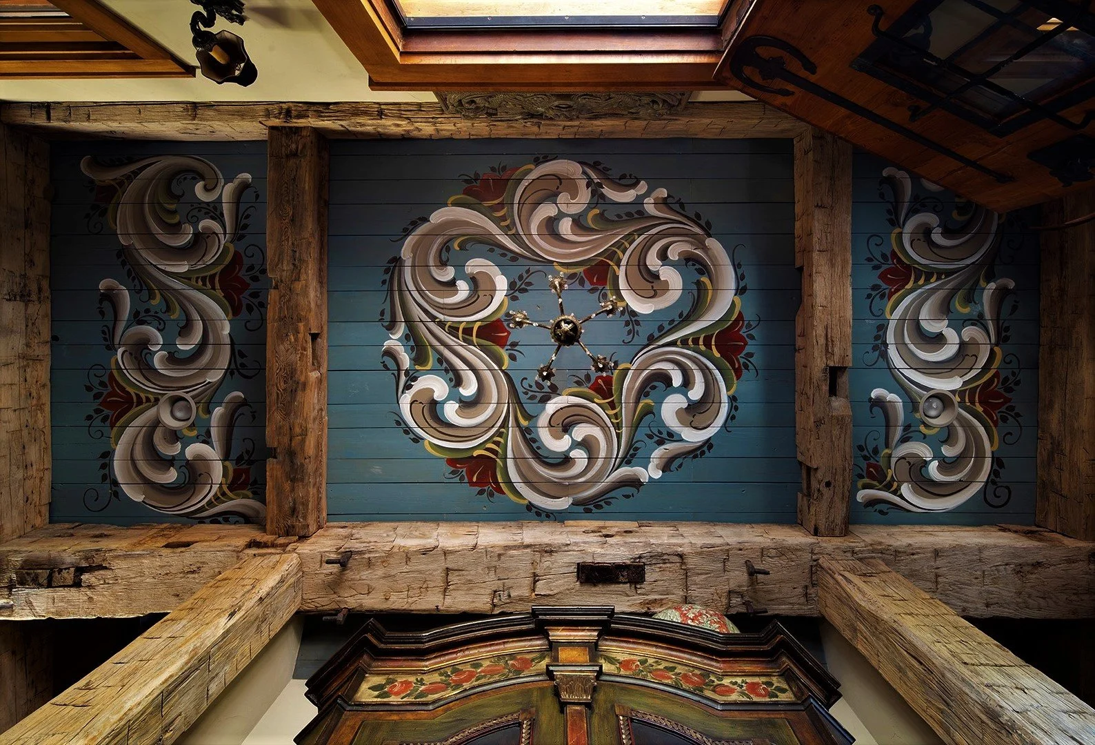 Ceiling with a decorative mural, featuring swirling patterns in beige, brown, red, and black on a blue background, with a small chandelier at the center and exposed wooden beams.