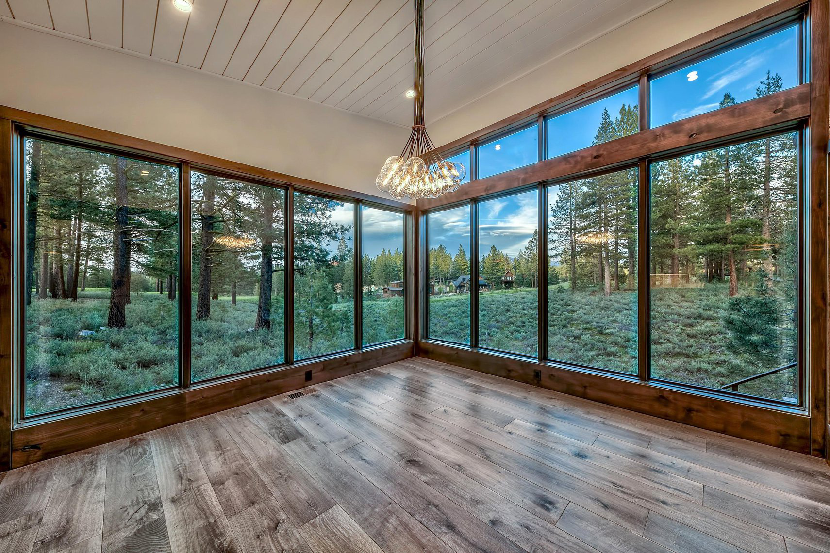 Empty room with large glass windows showing a forest landscape outside, wood flooring, and a modern chandelier hanging from the ceiling.