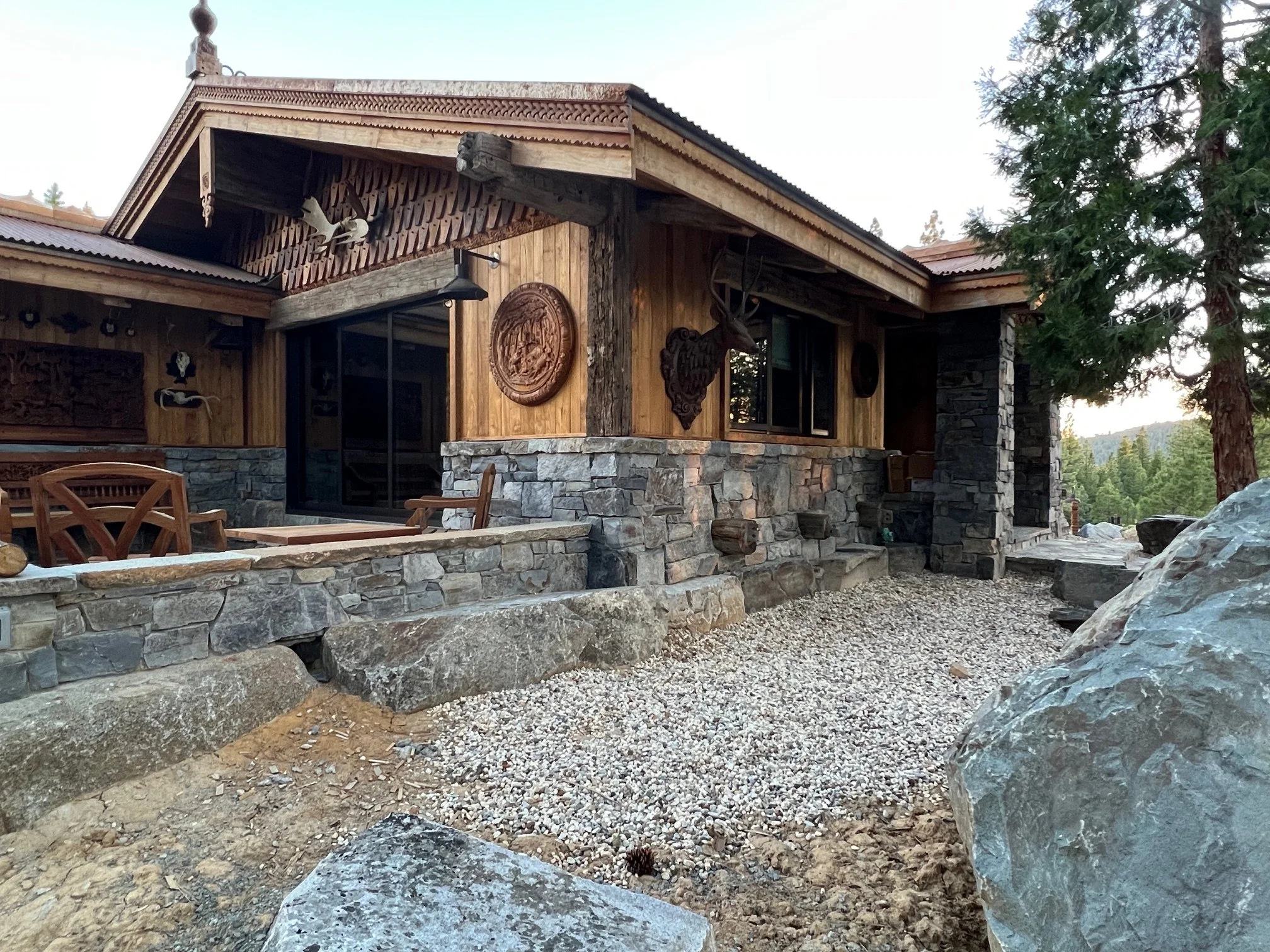 A rustic mountain house with stone and wood exterior, large rocks in front, surrounded by pine trees, with decorative carvings on the wall.
