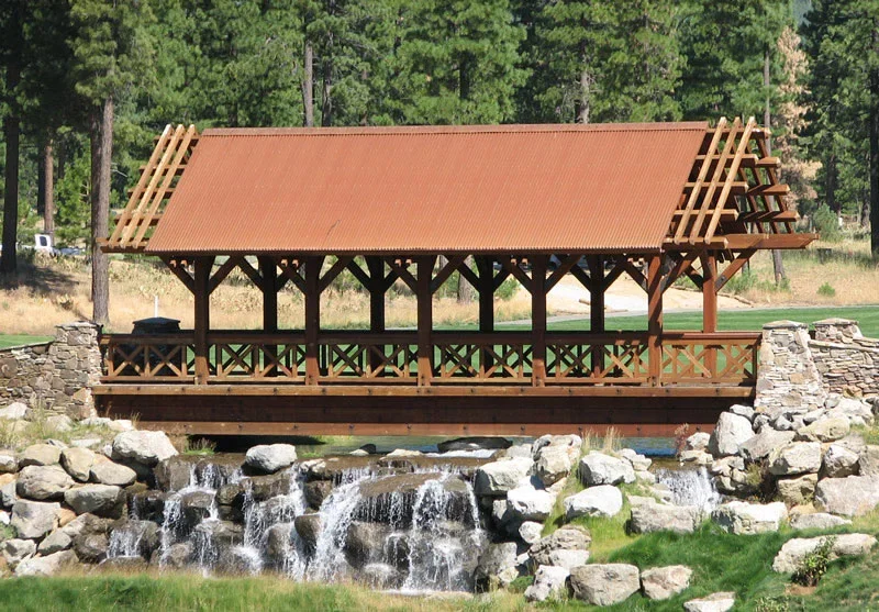 A wooden covered bridge with a red roof, spanning over a small waterfall and rocky stream, surrounded by green trees and grass.
