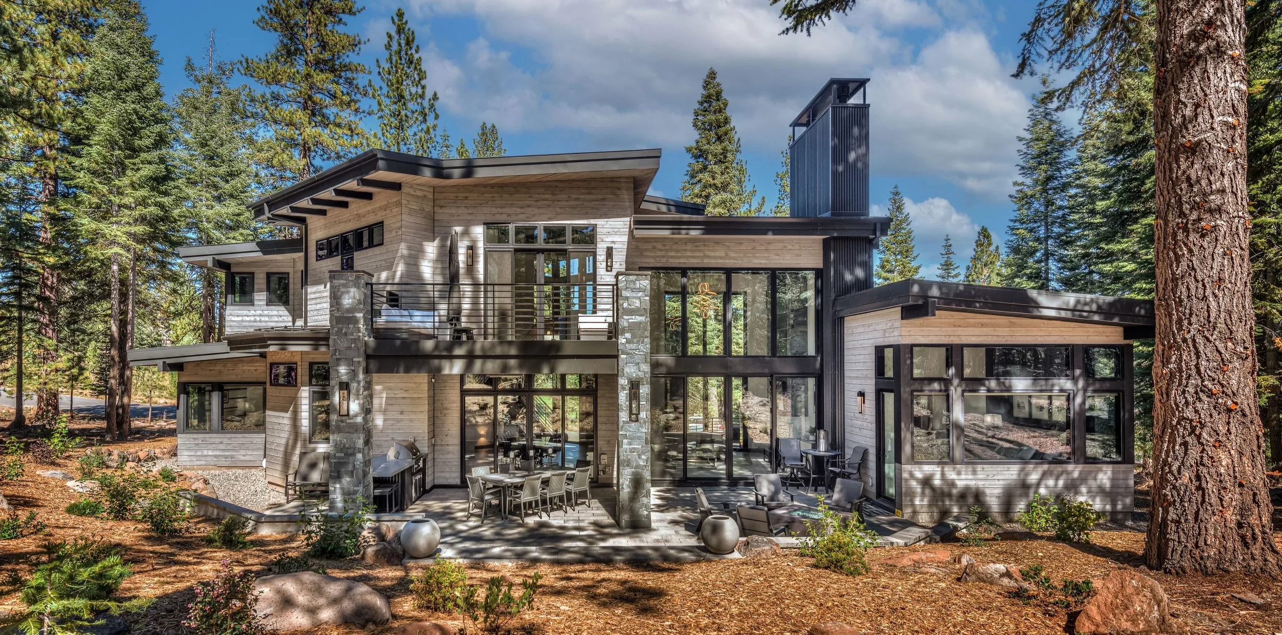 Modern multi-story house with large glass windows, wood and stone exterior, surrounded by tall pine trees, outdoor seating area, and patio under a partly cloudy sky.