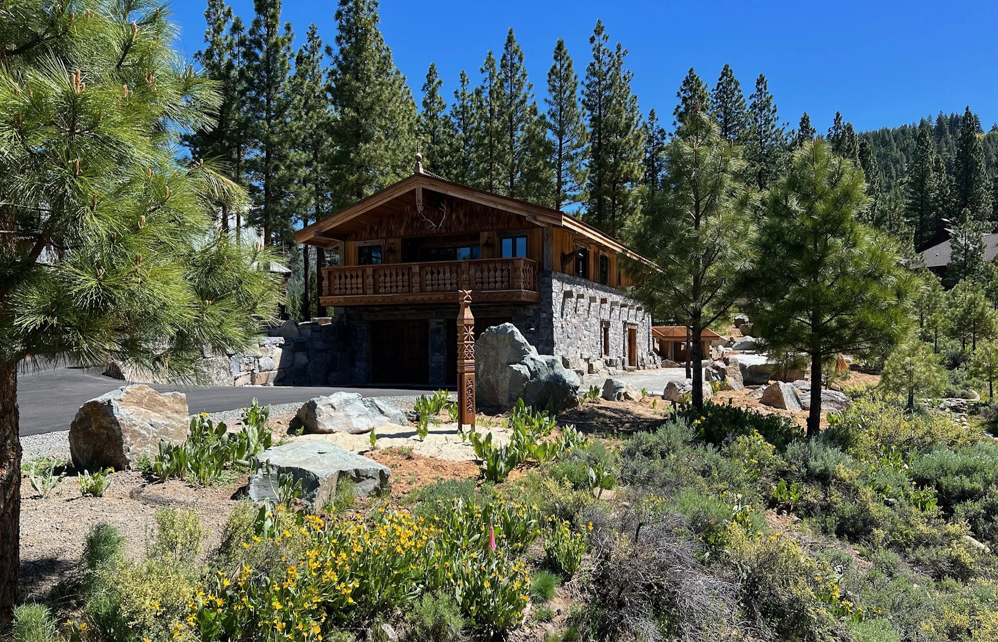 A rustic mountain house with a stone lower level and wooden upper level, surrounded by pine trees, rocks, and flowering plants, under a clear blue sky.