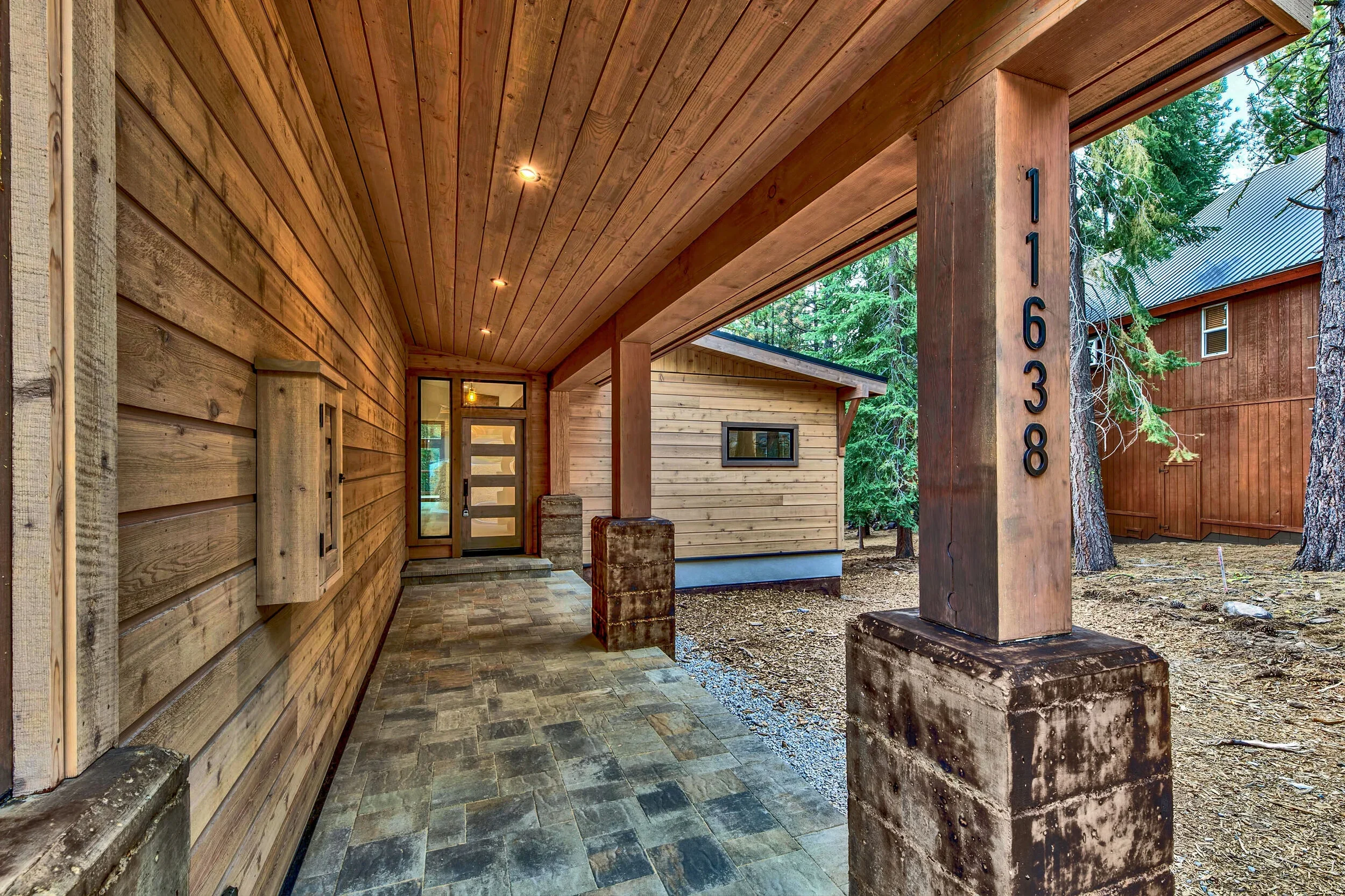 Close-up of a house's front porch with wooden siding, a supporting pillar with the address numbers 11638, and a stone-tiled floor. In the background, surrounding trees and part of another wooden building are visible.