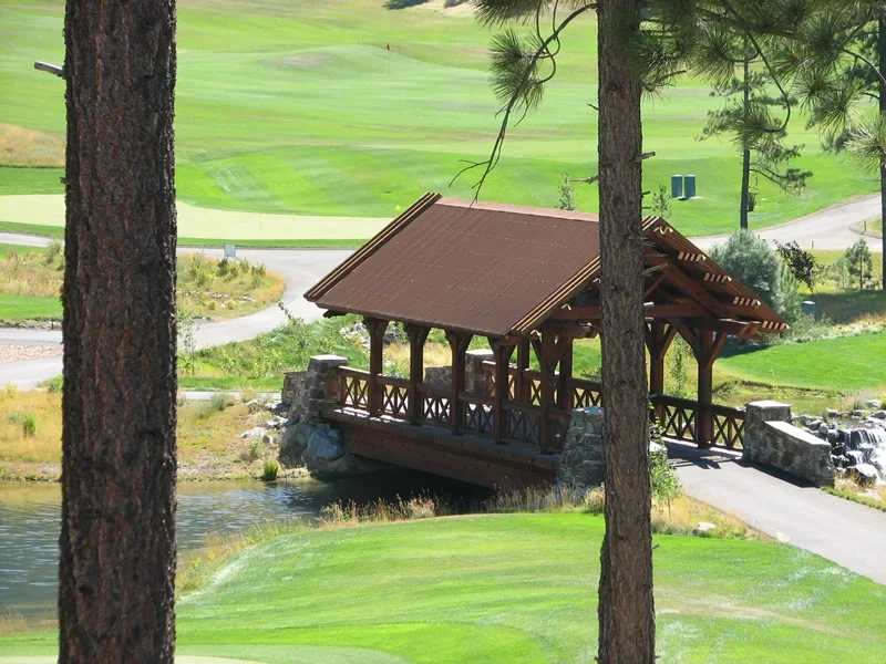 A wooden bridge with a roof crosses over a small pond at a golf course, with green grass and trees in the background.