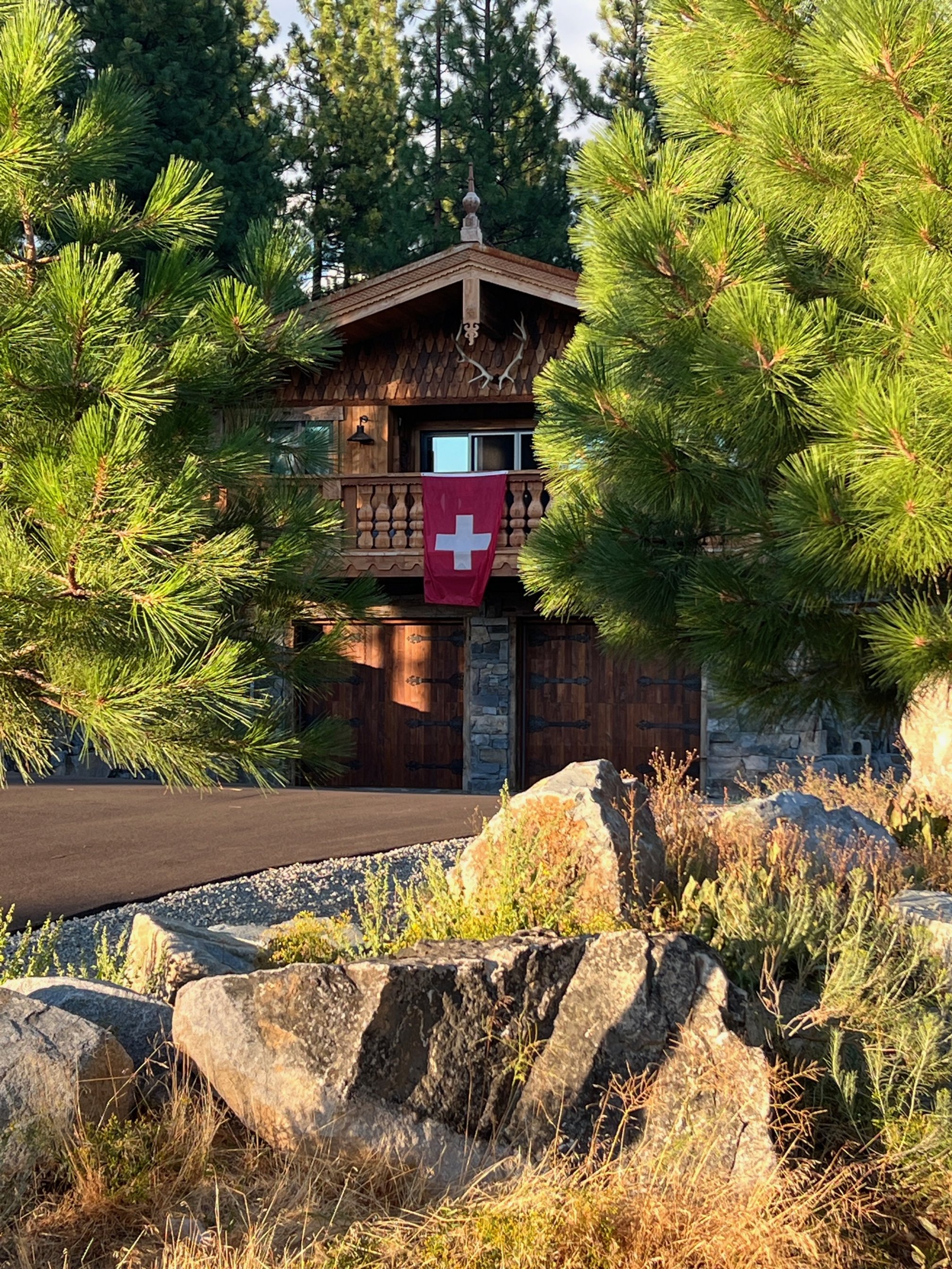 A wooden cabin with a Swiss flag hanging from the balcony, surrounded by green pine trees and rocks, with sunlight illuminating the scene.