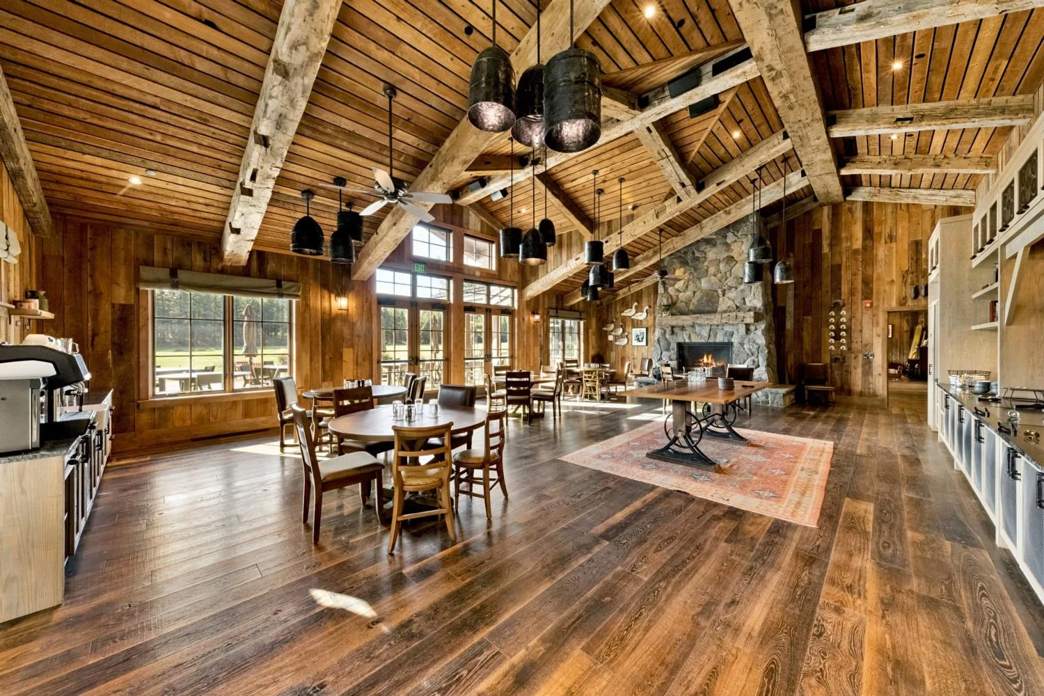 Large lodge dining hall with round wood tables, a stone fireplace, vaulted plank ceiling, exposed timber trusses, varied black pendant lights, and expansive windows overlooking the grounds.