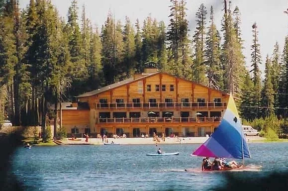 A lakeside scene with a large wooden lodge surrounded by pine trees, people on boats and paddleboards, and a sailboat with a colorful sail.
