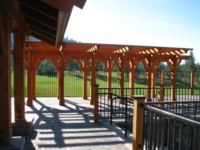 Empty outdoor wooden patio with lattice pergola and black metal railings, overlooking green fields and trees in the background.