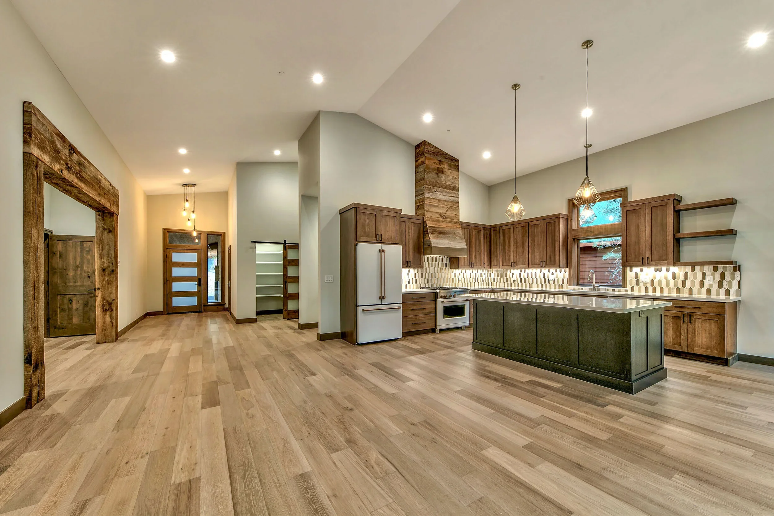 Open concept kitchen with wooden cabinets, modern backsplash, kitchen island, and pendant lighting, with wooden flooring and doorways leading to other rooms.