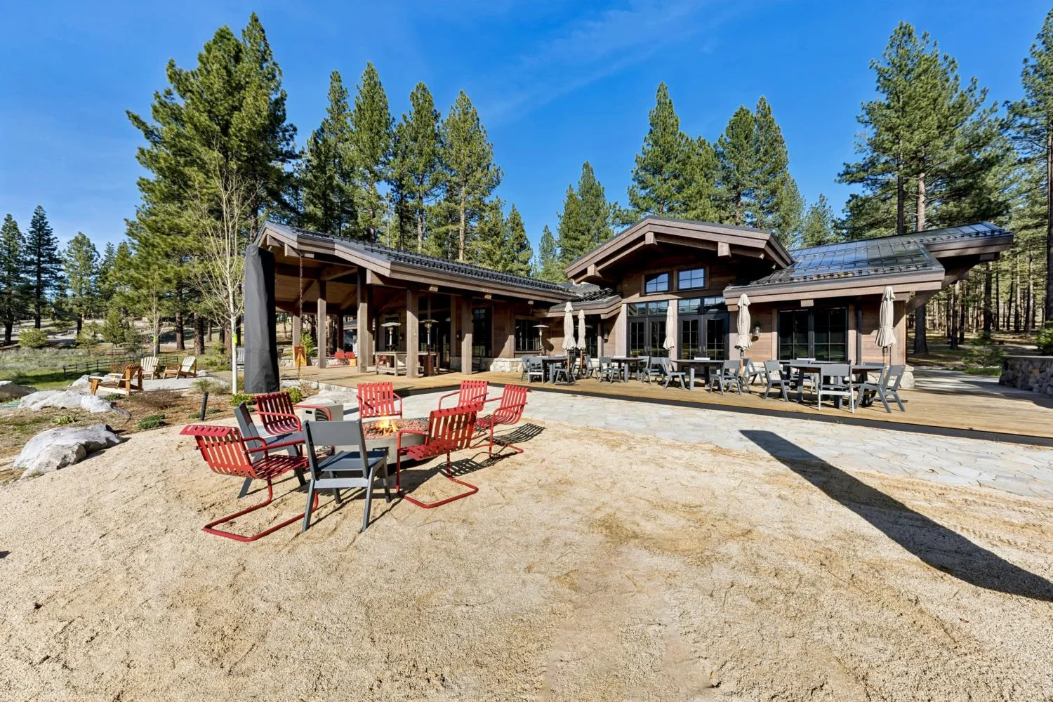 Modern mountain lodge with a broad wood deck, outdoor dining tables, umbrellas, a covered patio, and a fire pit surrounded by chairs beneath tall pine trees.