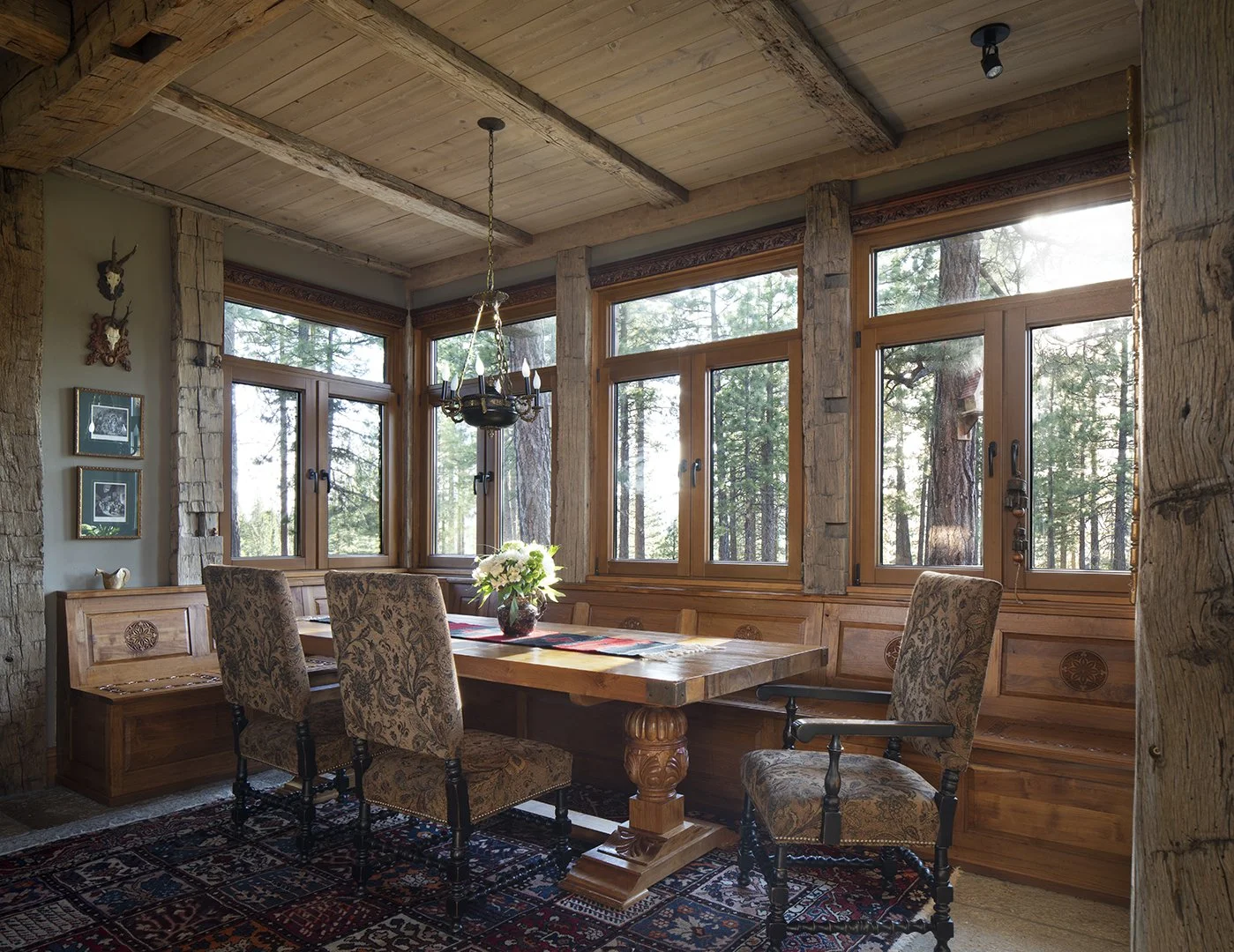 A cozy dining room with rustic wood decor, large windows overlooking a wooded forest, a wooden table with a flower arrangement, patterned upholstered chairs, and a chandelier hanging from the ceiling.