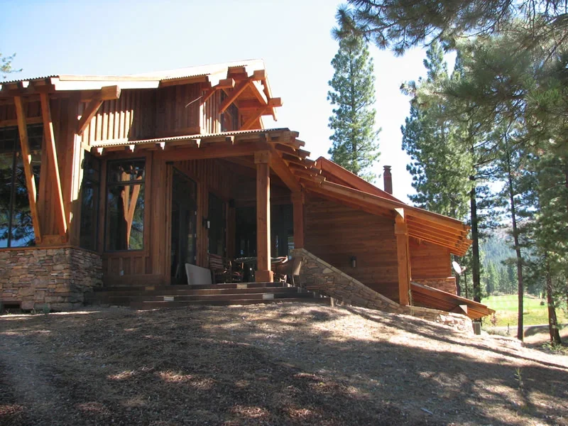 A rustic wooden house with a stone foundation, surrounded by tall pine trees in a forested area.