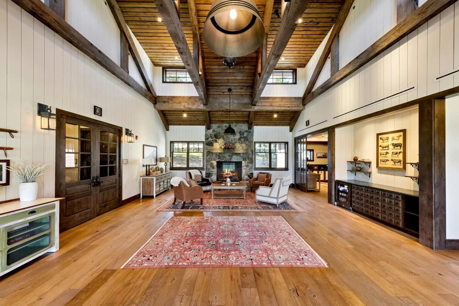 Spacious lodge lounge with a soaring wood-plank ceiling, exposed timber beams, natural wood flooring, patterned rugs, and a seating area centered around a stone fireplace.