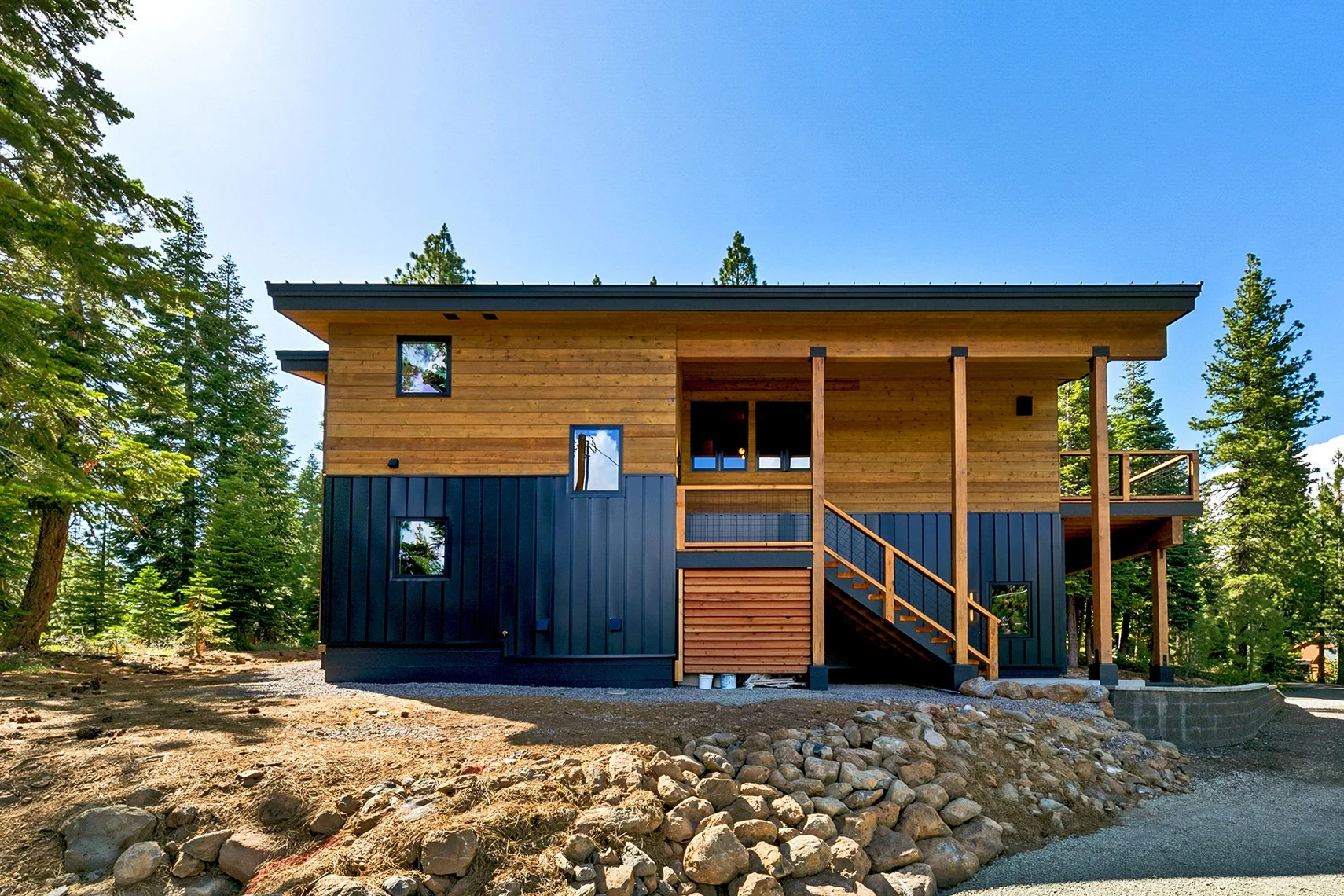 Modern mountain home exterior featuring natural wood siding, black vertical metal panel accents, exposed timber posts, an elevated front entry staircase, and a covered entryway deck surrounded by pine trees.