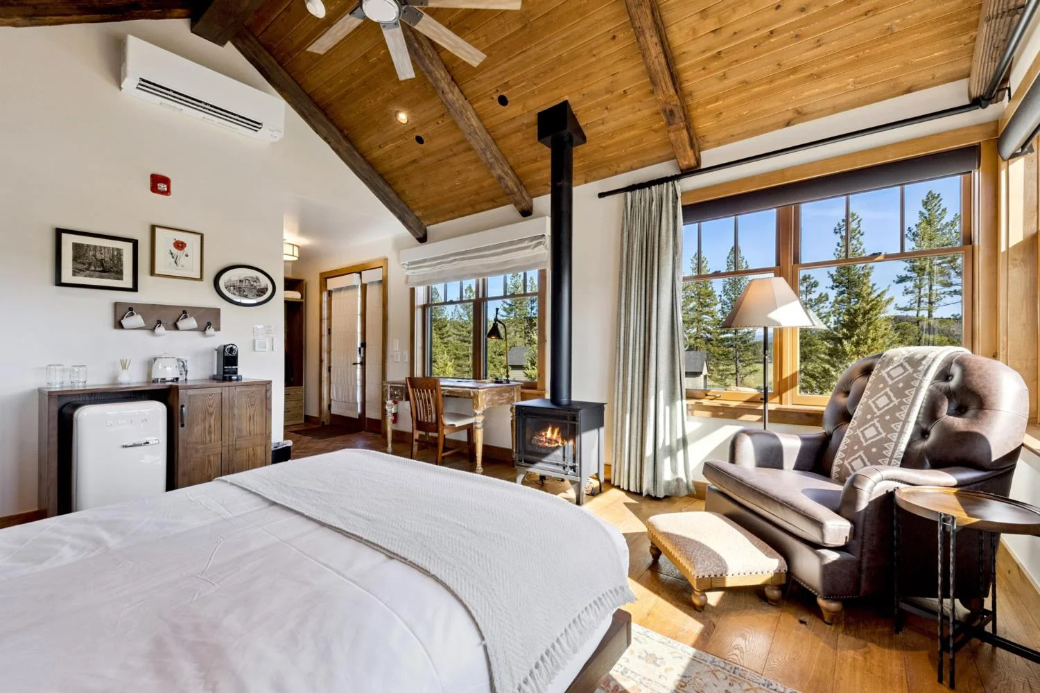 Private cabin bedroom with a vaulted wood ceiling, exposed beams, a freestanding stove with a black chimney pipe, leather reading chair, writing desk, coffee station, and large forest-facing windows.