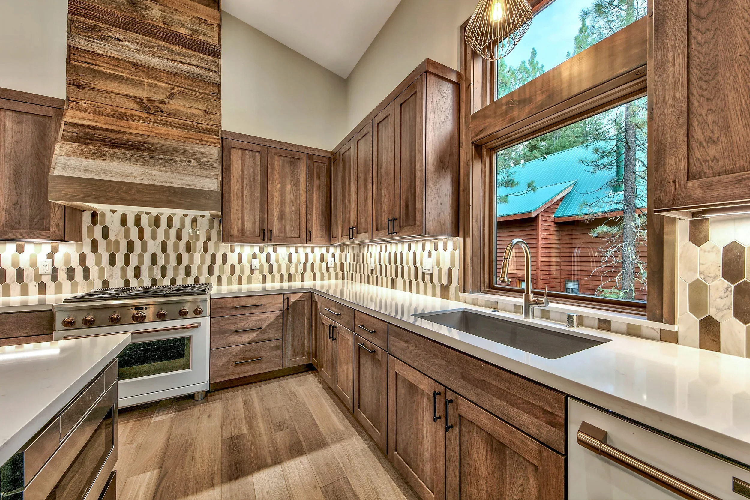 Kitchen with wooden cabinets, white countertops, hexagonal tile backsplash, large window revealing outdoor trees and a building with a green roof.