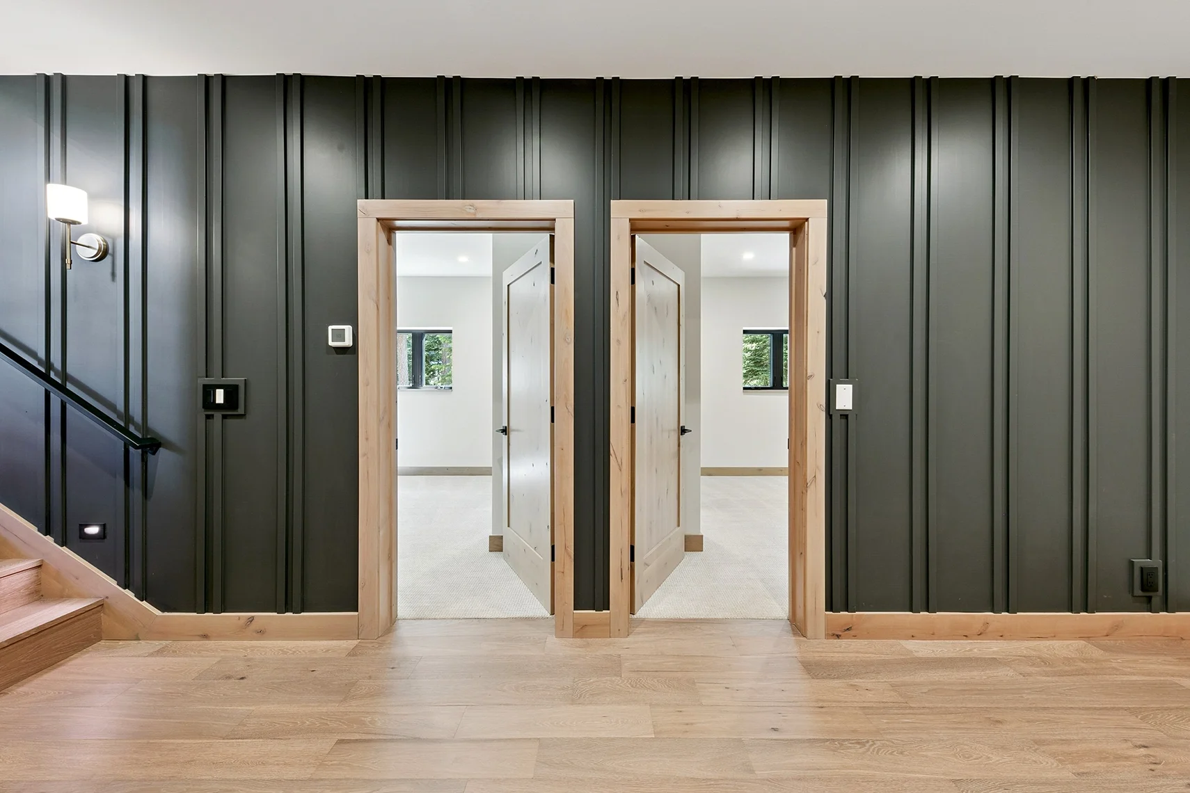 Modern hallway with light wood flooring, two open mirrored bedroom doorways, a dark green wood-paneled wall, and stairs leading to the upper level.