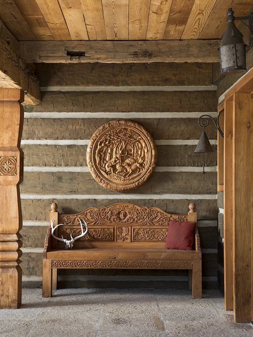 Rustic wooden interior with carved wooden bench, red cushion, antler decor, and circular wood carving on the wall, surrounded by stone and wood walls.