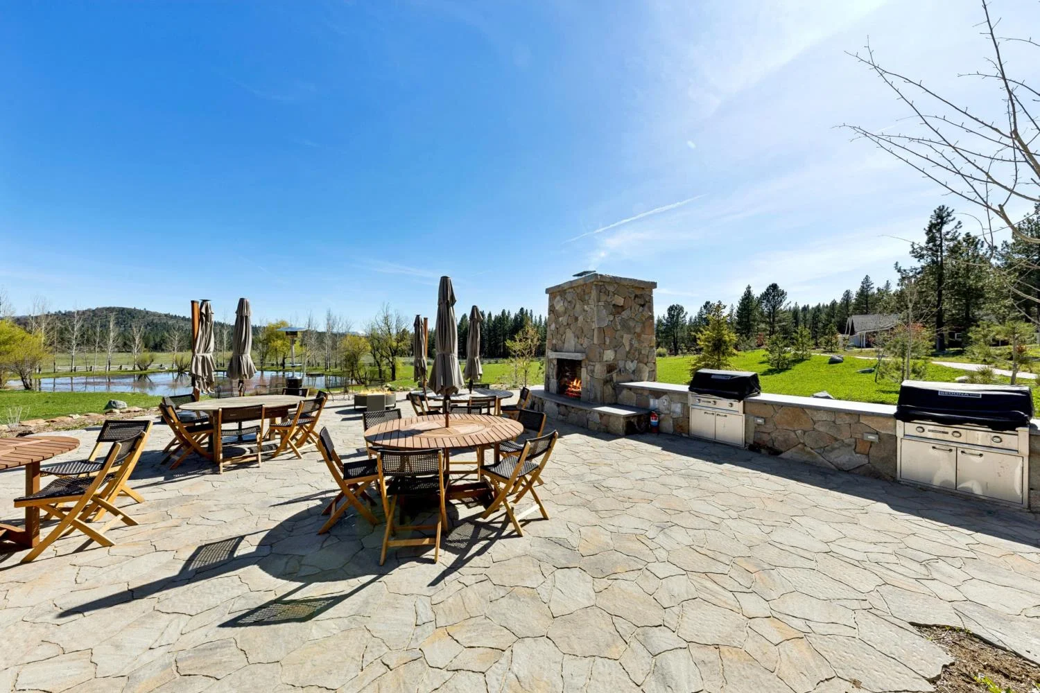 Large stone dining patio with round tables, an outdoor fireplace, built-in grills, and views of a pond, green meadow, and surrounding pine trees.