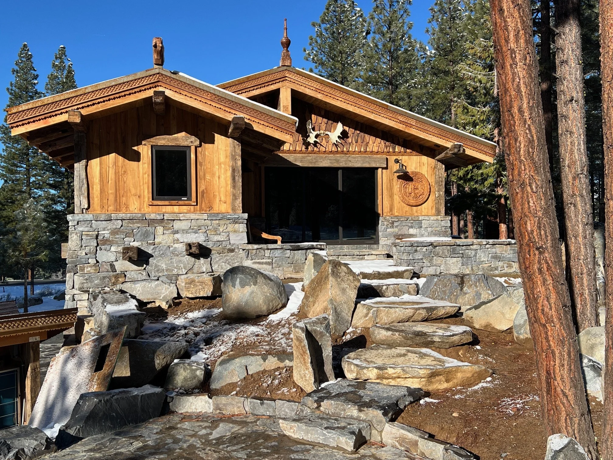 A wooden cabin with stone foundation, surrounded by tall trees, with rocks and snow on the ground in front.