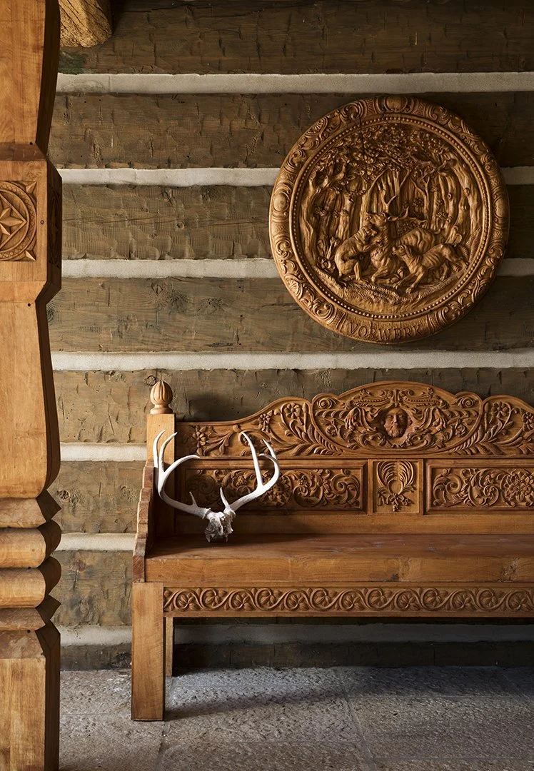 Wooden bench with intricate carvings and a mounted deer skull with antlers on the left side. A carved round plaque depicting a hunting scene hangs above the bench. The background features a rustic wall made of horizontal logs.
