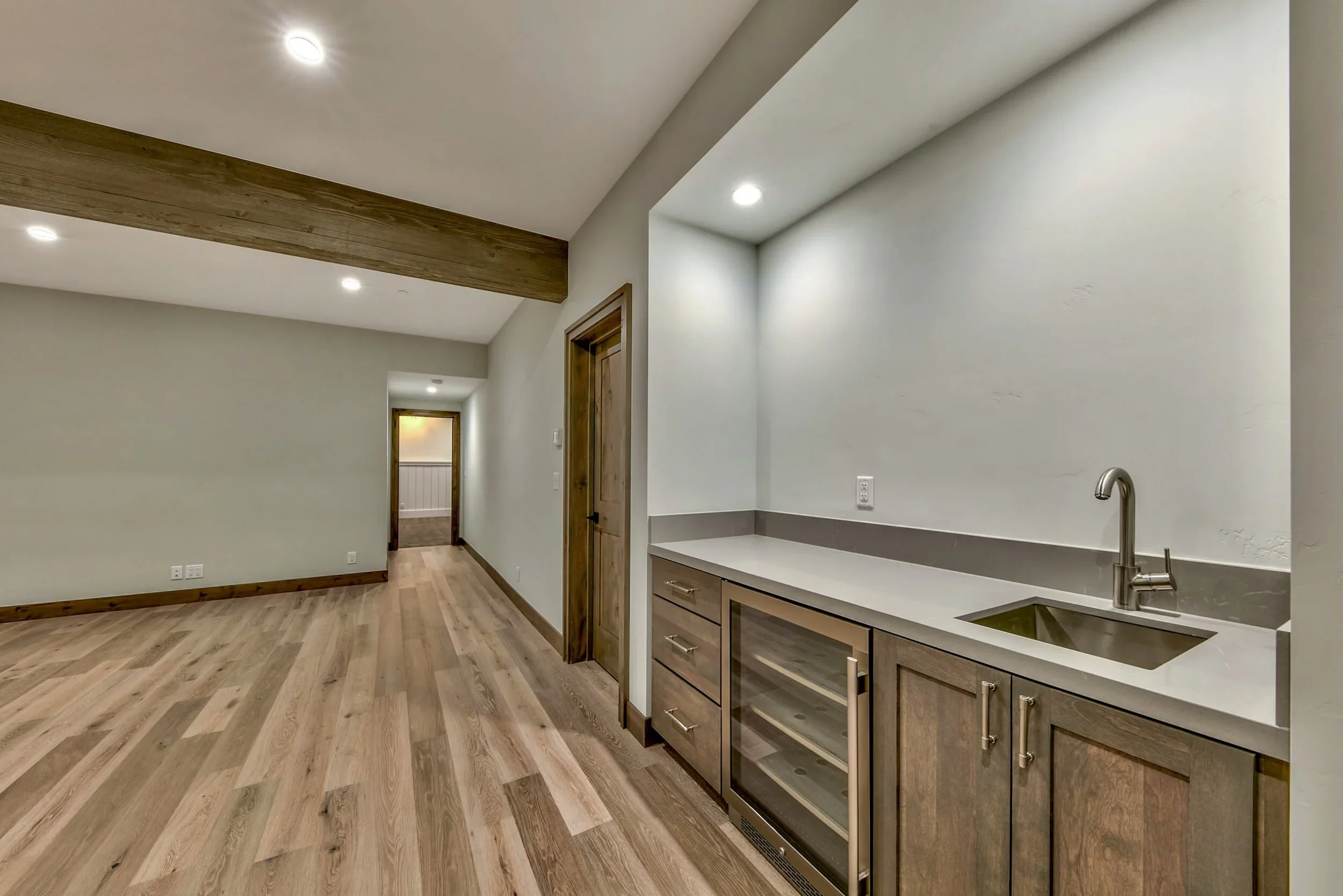 Empty room with light wood flooring, white walls, a small kitchenette with a sink, wine fridge, and drawers, and a hallway leading to a space with a white railing.