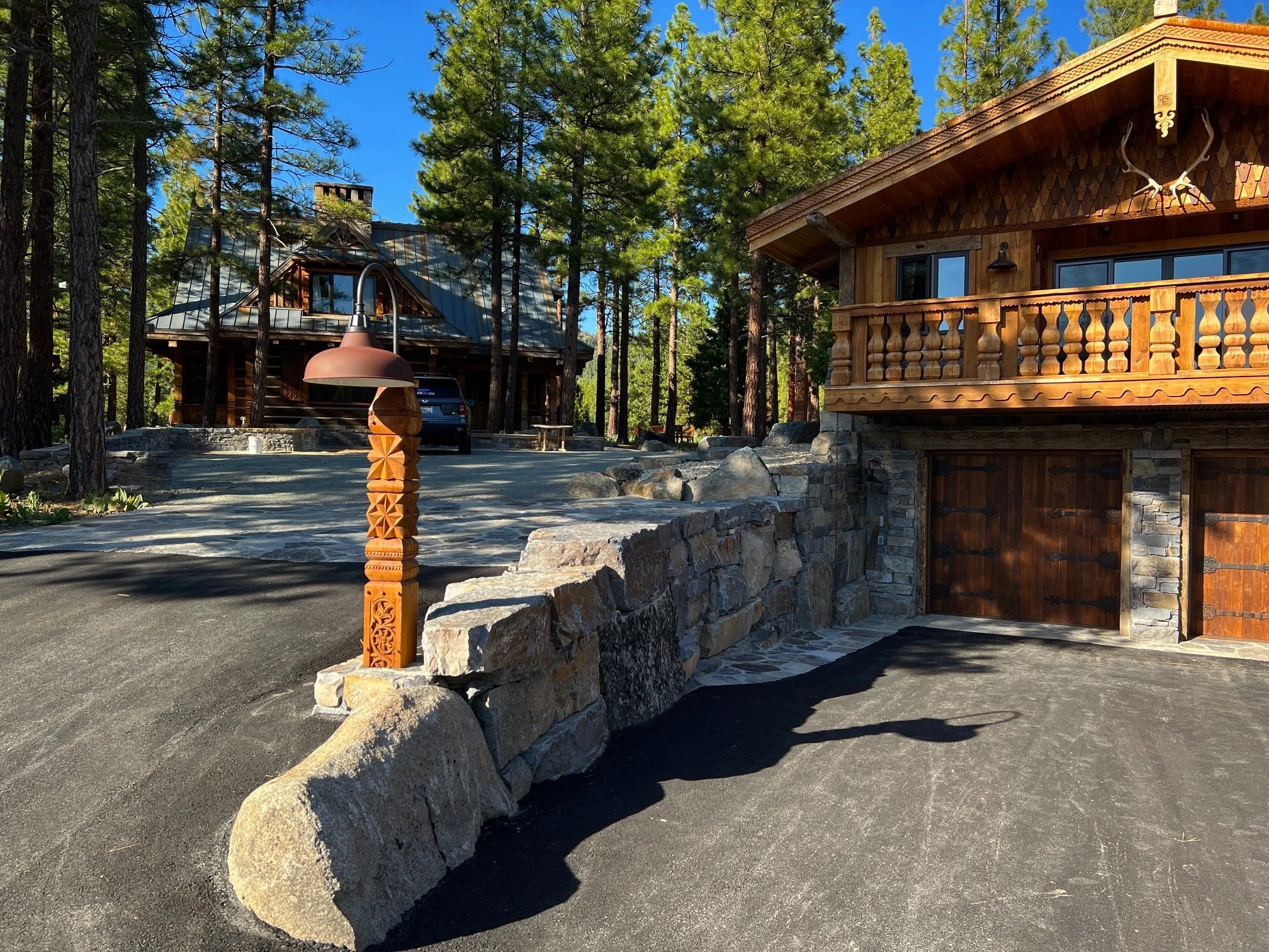 A rustic wooden house in a forested area with a stone driveway, a vintage-style lamp post, and deer antlers mounted on the upper wall.