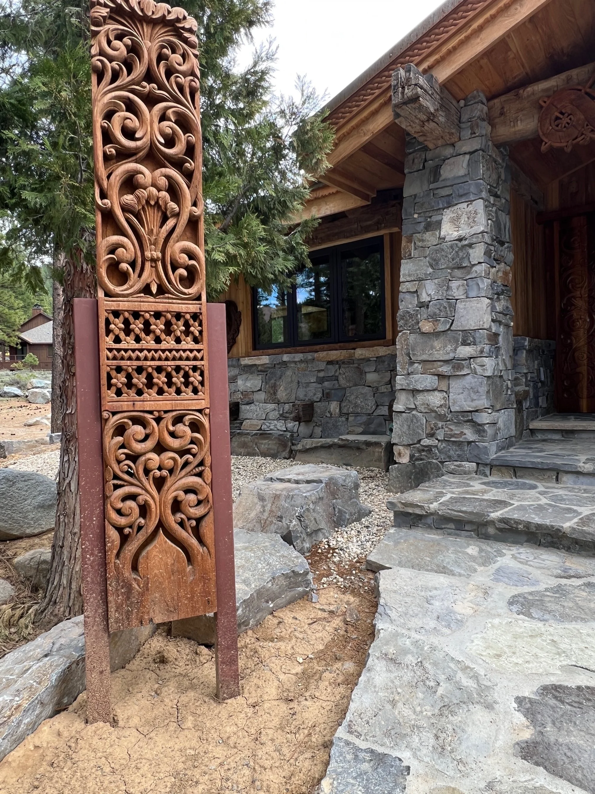 A decorative wooden post with intricate carvings standing next to a stone and wood house with large stones on the pathway.