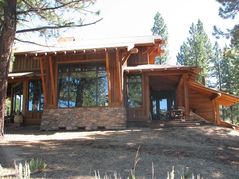 A modern house with a wooden exterior and large glass windows, situated in a pine forest.