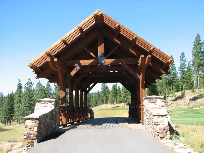 A wooden covered bridge with stone supports on a paved road in a scenic, rural area with green grass and pine trees.