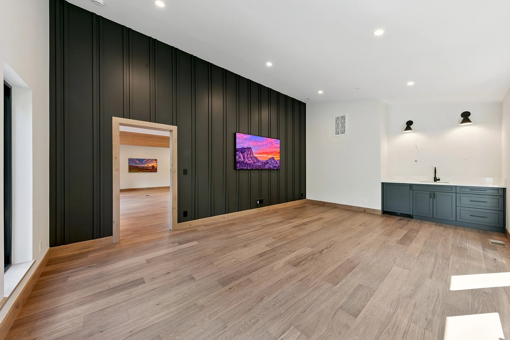 Modern living space with built-in cabinetry, white walls, a black wood-paneled accent wall with a TV, and a doorway leading to an additional living area.