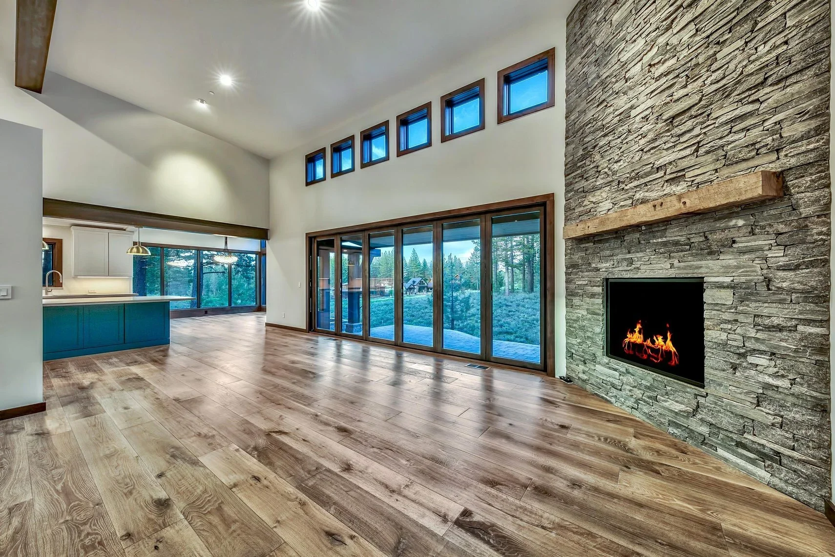 Open-concept living room with wood flooring, stone fireplace, large sliding glass doors, high ceiling with small upper windows, and visible kitchen in the background.