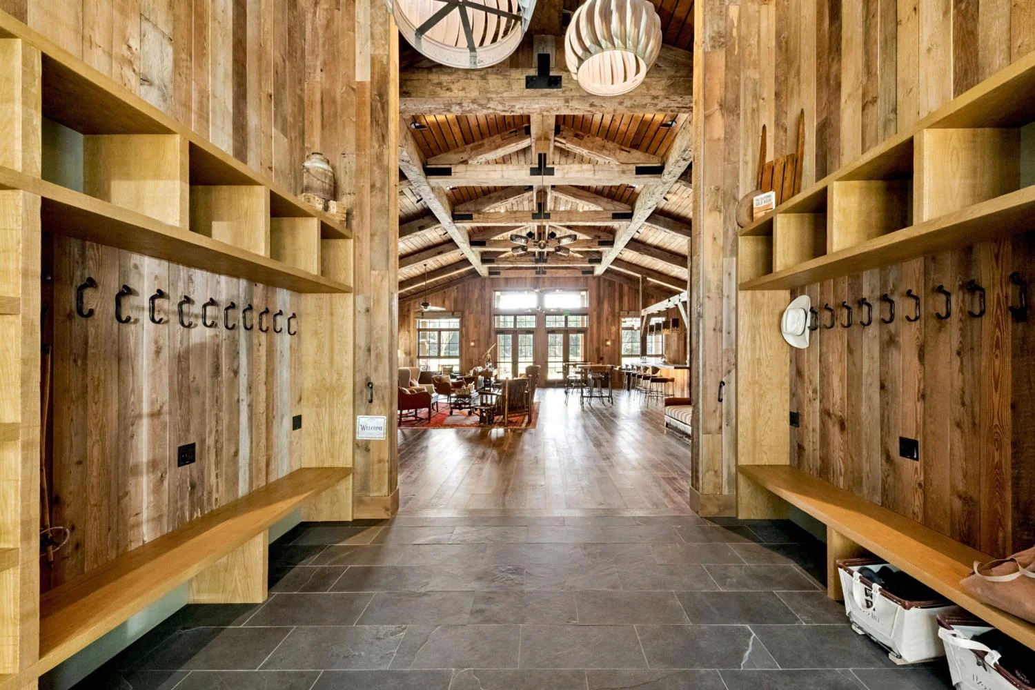 Lodge entry with reclaimed wood walls, built-in benches, open cubbies, black coat hooks, slate tile flooring, and a vaulted timber-framed lounge beyond.