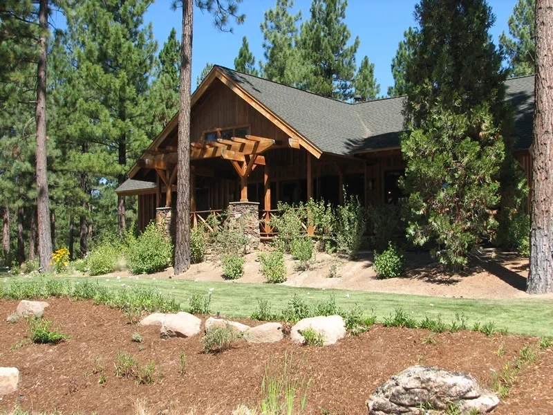 A wooden house surrounded by tall pine trees with a landscaped front yard featuring small shrubs and flowers.