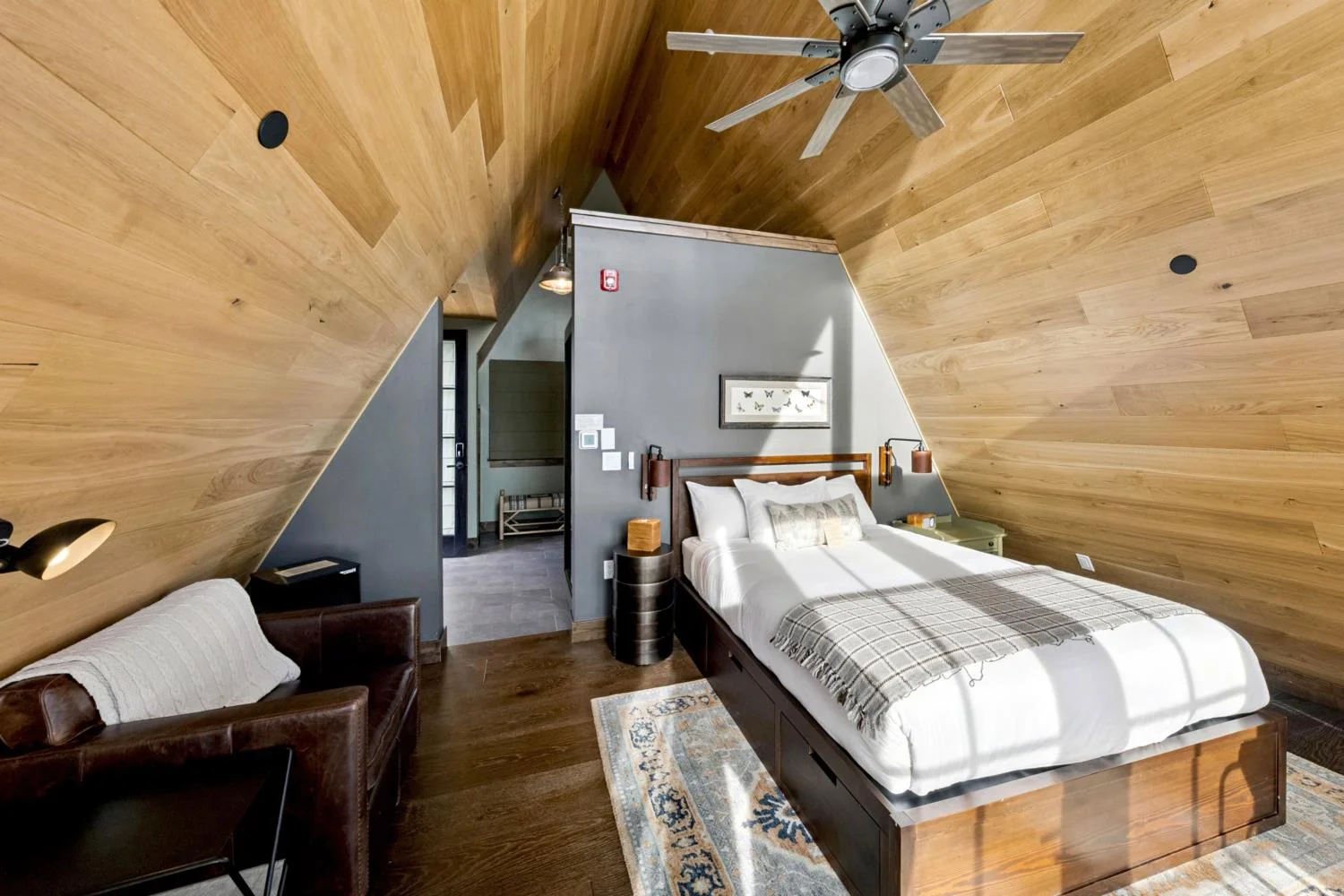 A-frame cabin bedroom with sloped wood-paneled walls, dark wood flooring, a platform bed, leather loveseat, patterned rug, and ceiling fan.