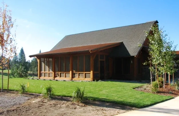 A house with a large sloped roof, wooden front porch, surrounded by a green lawn and young trees, under a clear blue sky.
