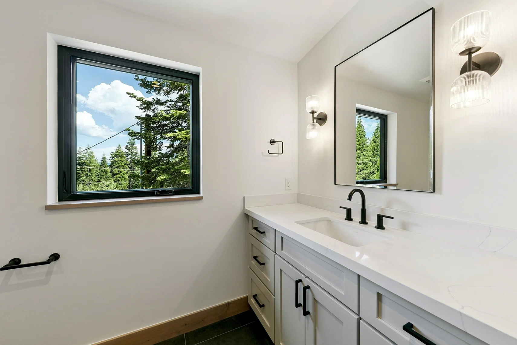 Modern white bathroom with black fixtures, a large mirror, and a window with a scenic outdoor view.