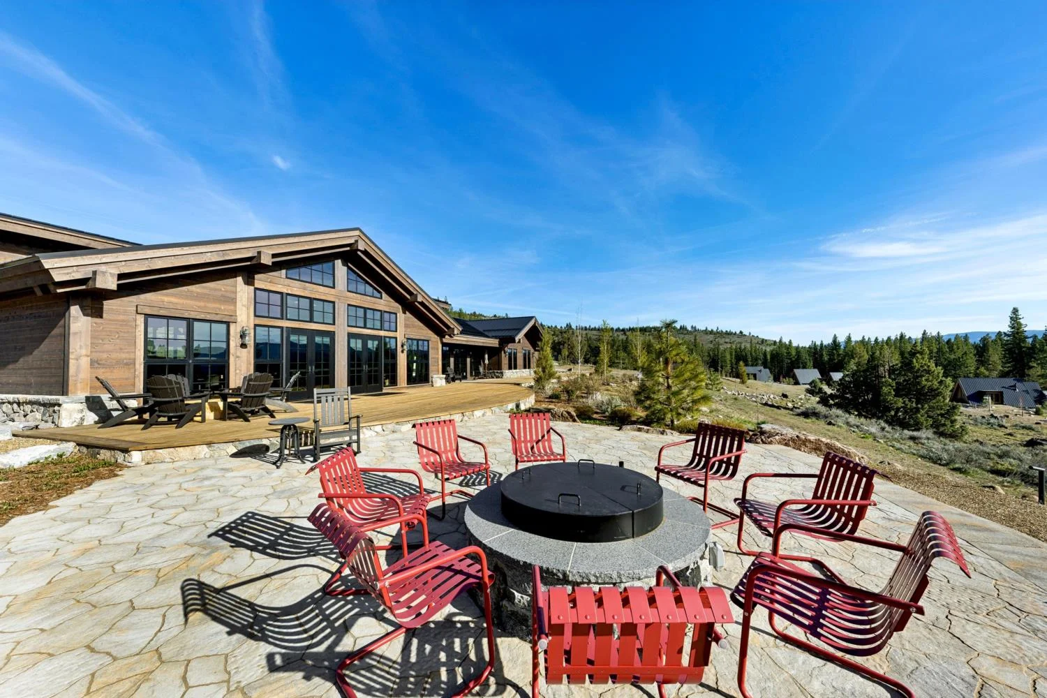 Stone patio with red chairs surrounding a circular fire pit beside a modern mountain lodge, overlooking pine trees, hillside cabins, and distant mountains beneath a blue sky.