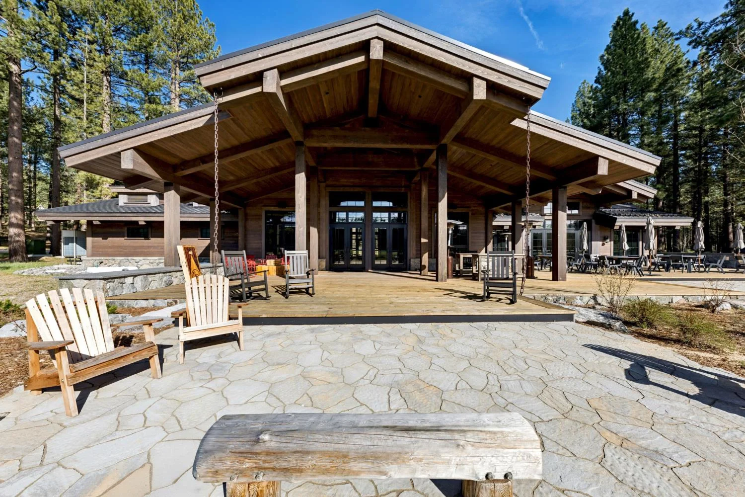 Modern mountain lodge with a broad timber-framed entry canopy, covered wood deck, hanging chairs, stone patio, and outdoor seating surrounded by pine trees.