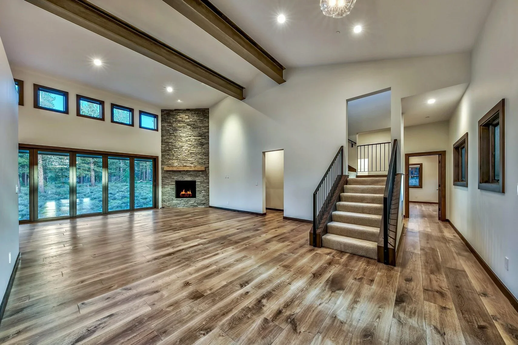 Empty living room with hardwood floors, large windows, and a stone fireplace with a wooden mantle.