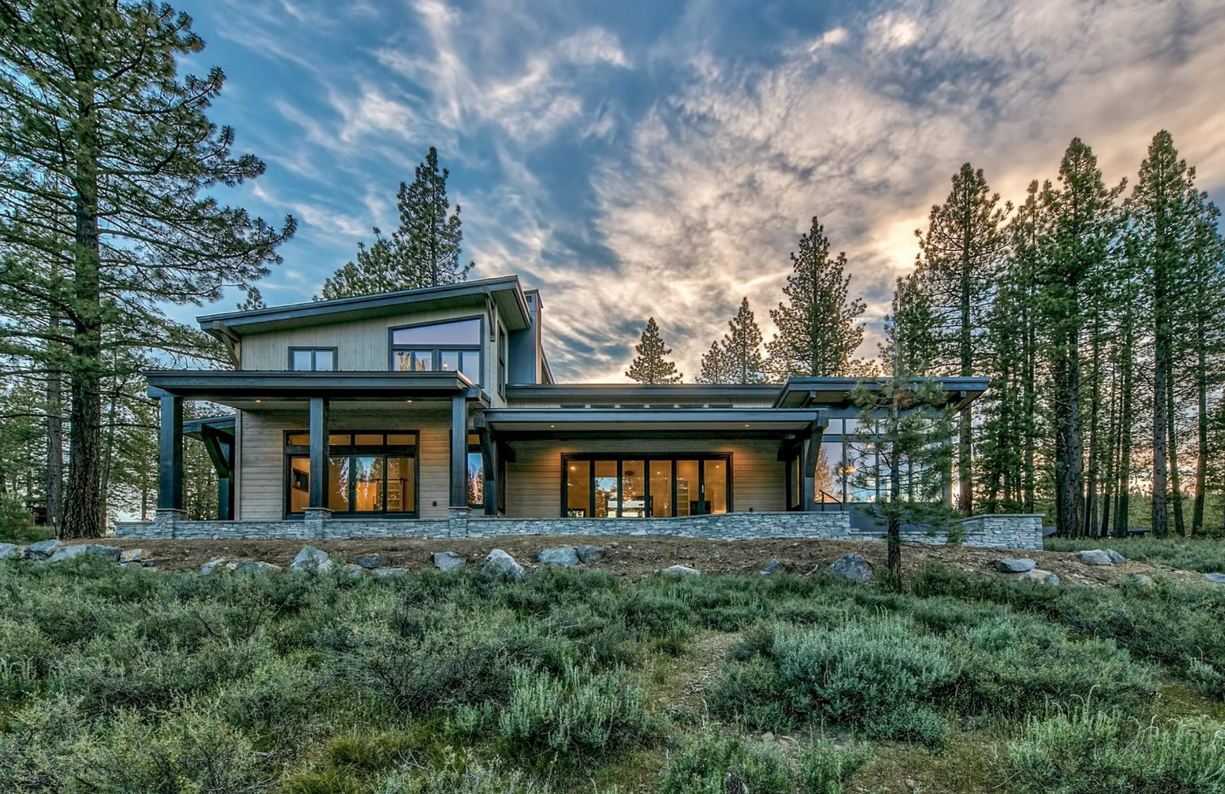 Modern two-story house with large windows, set among tall pine trees, under a partly cloudy sky at sunset.