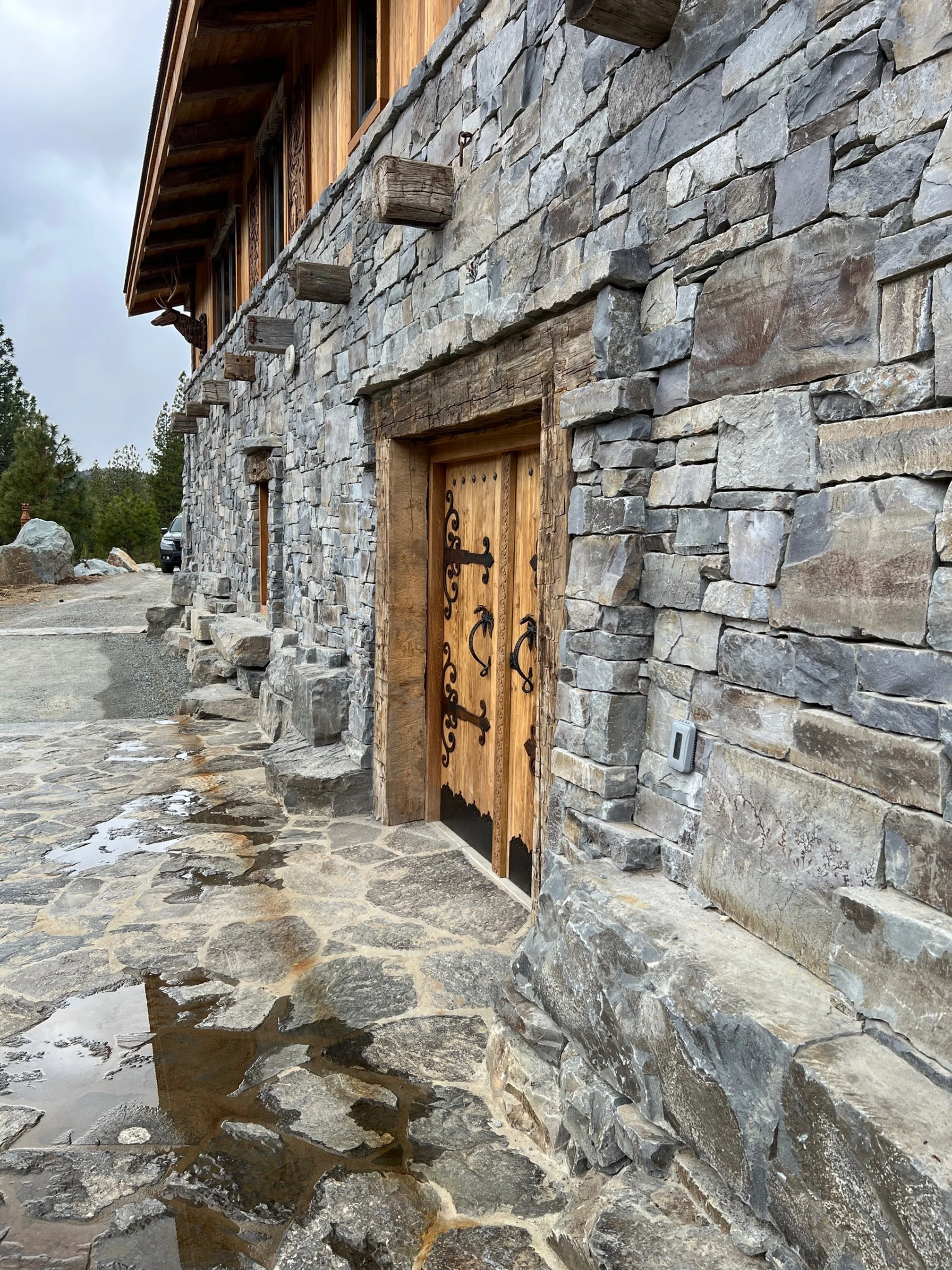 Stone house with wooden door featuring decorative black iron hinges and handles, outdoor stone pathway, and stone wall exterior with small window and overhanging roof.