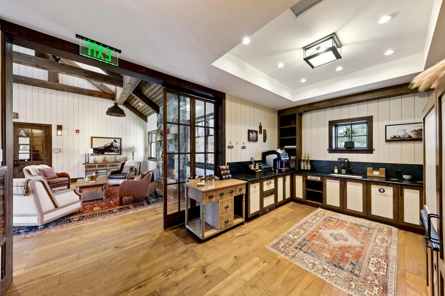 Lodge refreshment area with a coffee station, black countertops, wood-and-white cabinetry, a small sink, natural wood flooring, a square patterned rug, and glass doors opening to a fireside lounge.