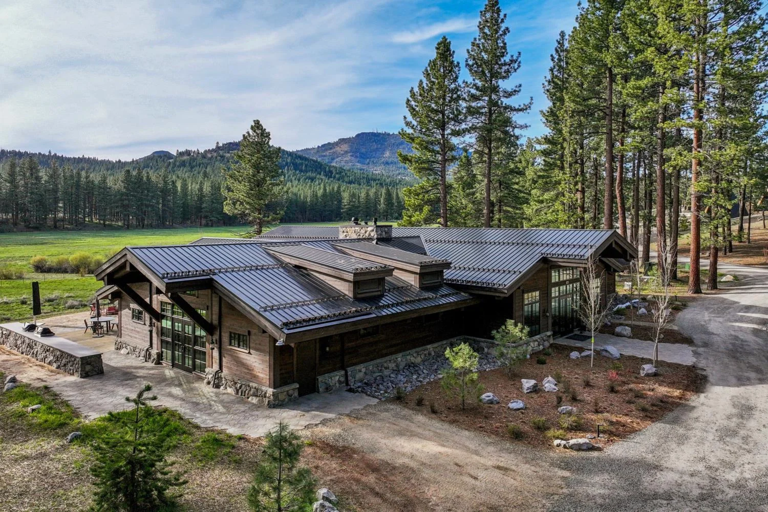 Modern mountain lodge with natural wood siding, stone detailing, a dark metal roof, and expansive windows overlooking a pine forest, green meadow, and distant mountains.