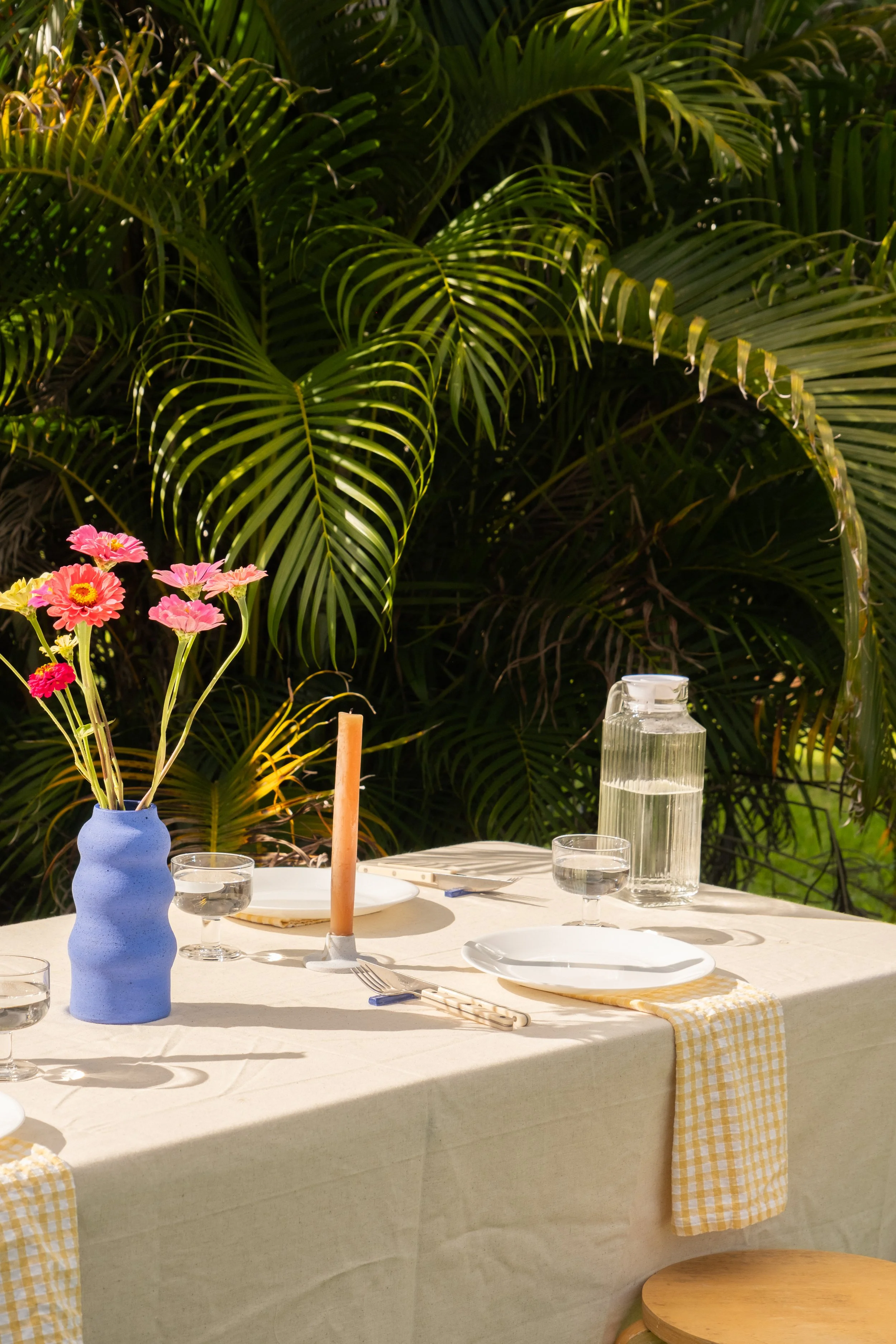 An outdoor dining table set with white plates, a water pitcher, and glasses, decorated with a bouquet of pink and yellow flowers in a blue vase, and a pink candle, surrounded by lush green palm leaves.
