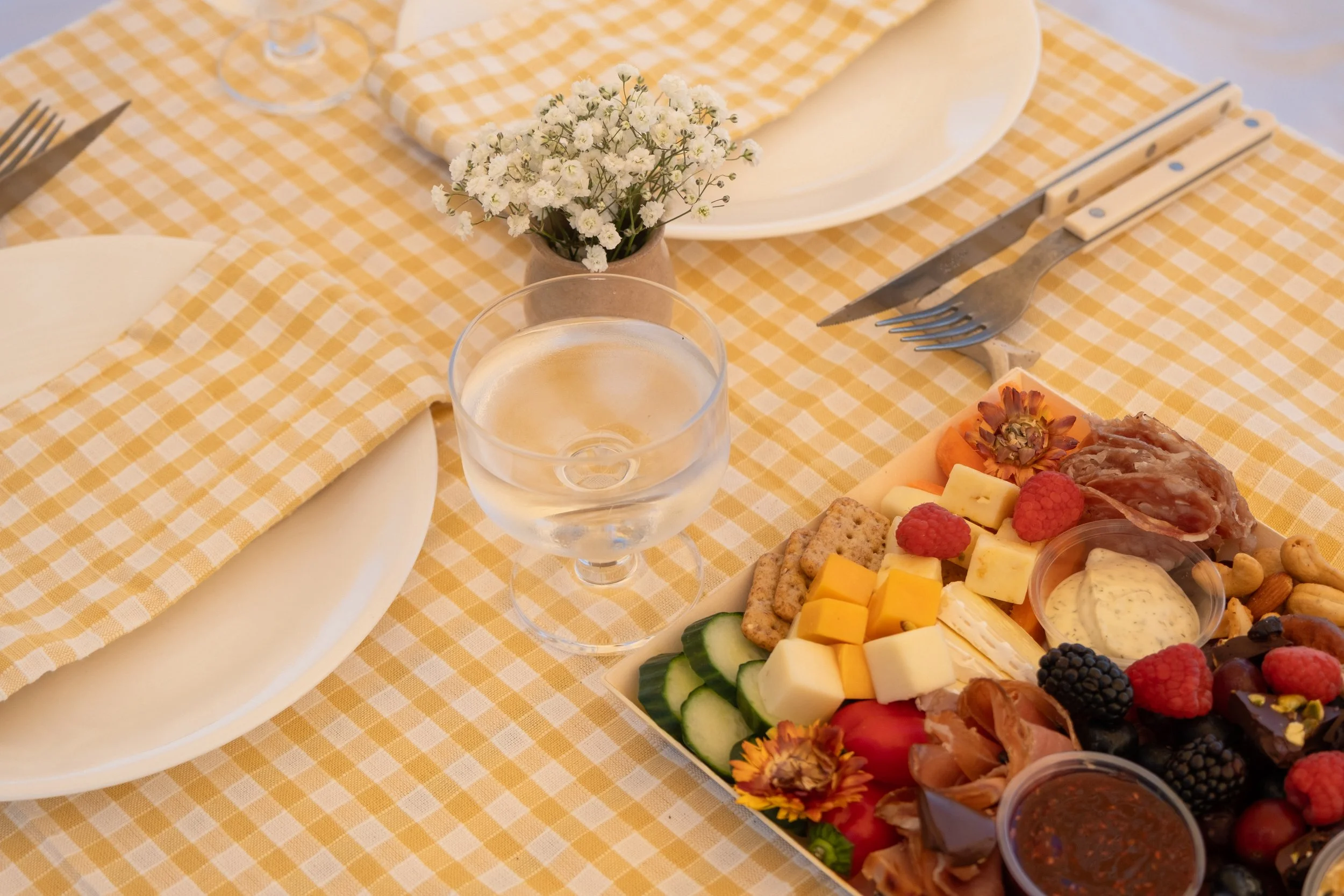 A table set with white plates, cutlery, a glass of water, a small vase of white flowers, and a cheese and fruit platter with various cheeses, sliced cucumbers, berries, nuts, and a small bowl of dipping sauce on a yellow-checkered tablecloth.
