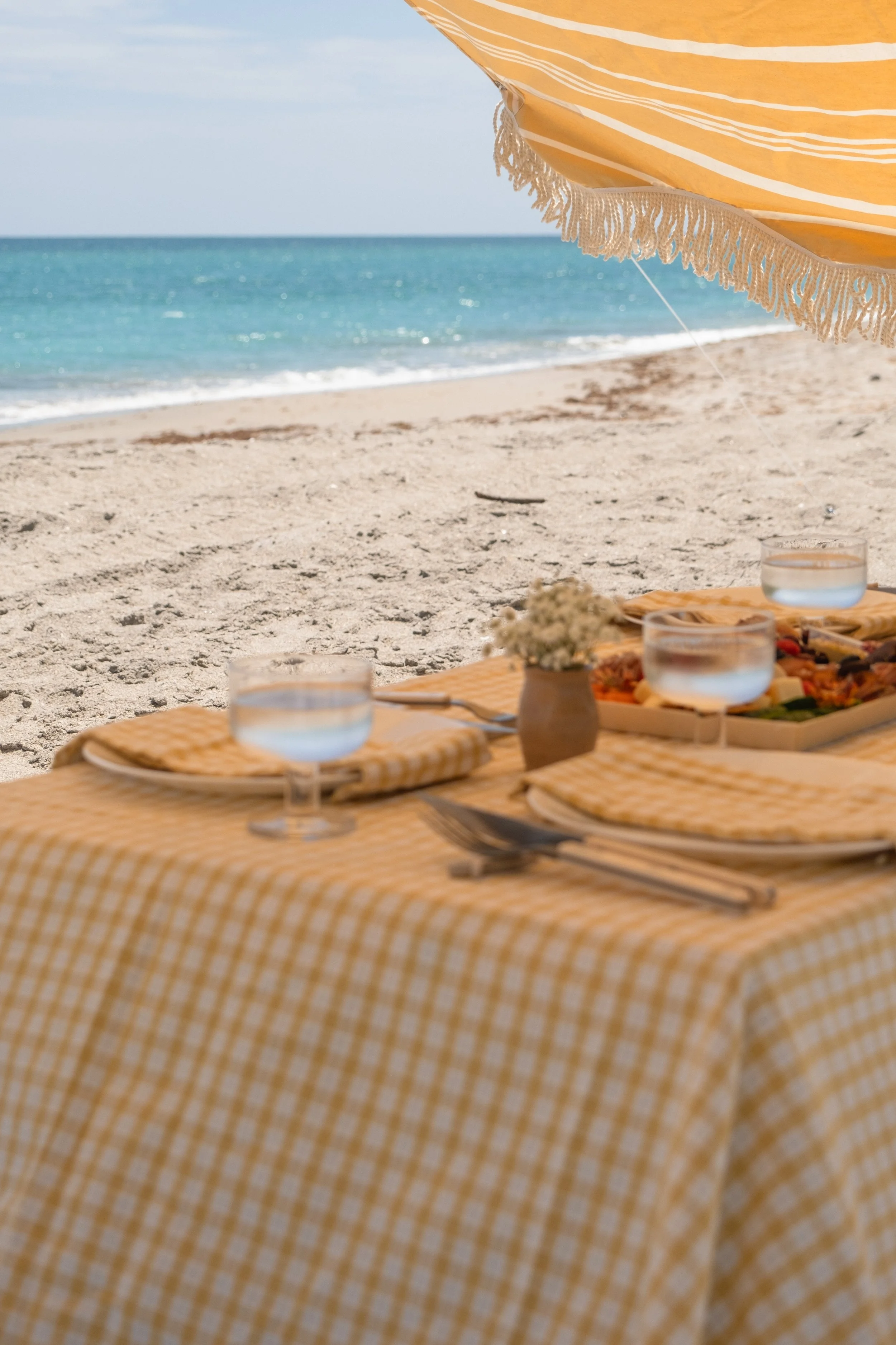 Beachside dining setup with a yellow checkered tablecloth, plates, glasses of water, and cutlery under a striped yellow and white umbrella, overlooking the sandy beach and blue ocean.