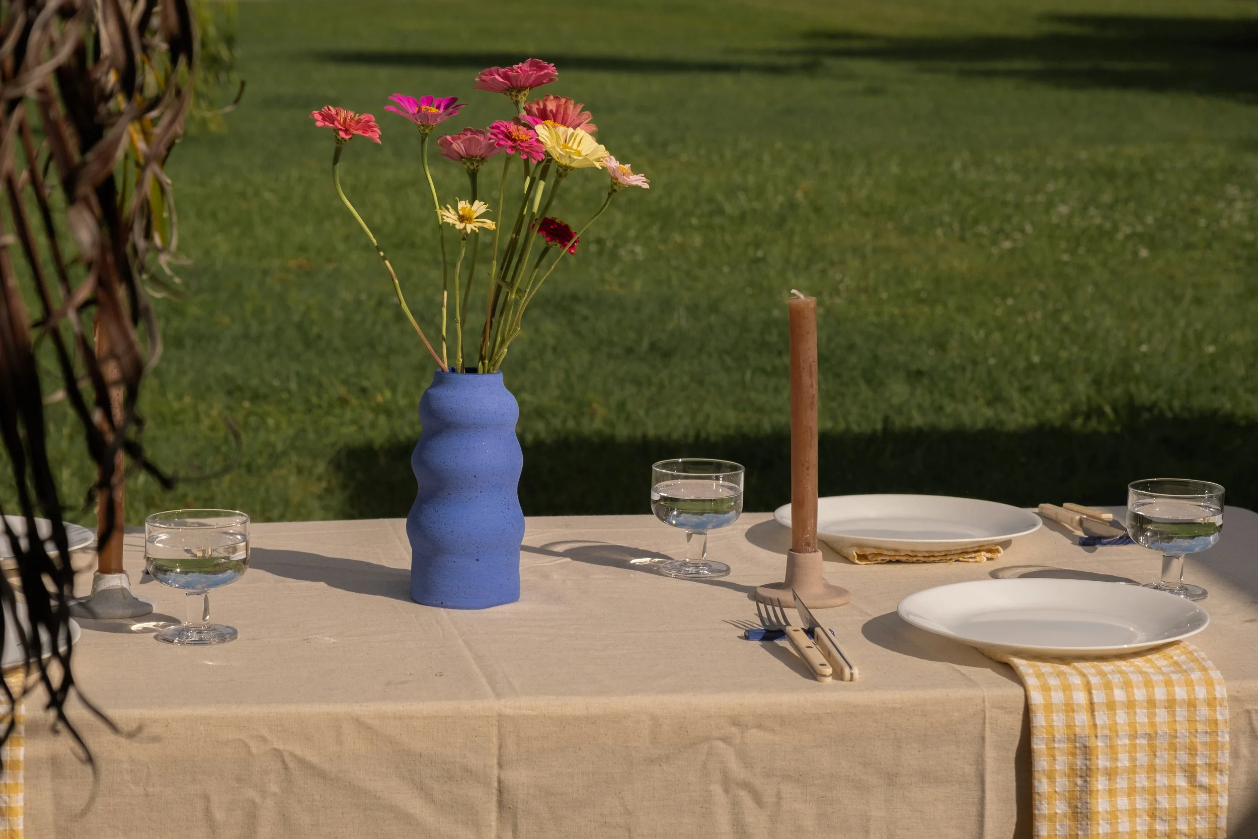 On an outdoor table, there is a blue vase with pink, yellow, and red flowers, a pink candle, three water glasses, white plates, beige napkins, and gold utensils, with a checkered cloth hanging from one plate, set on a beige tablecloth against a grass