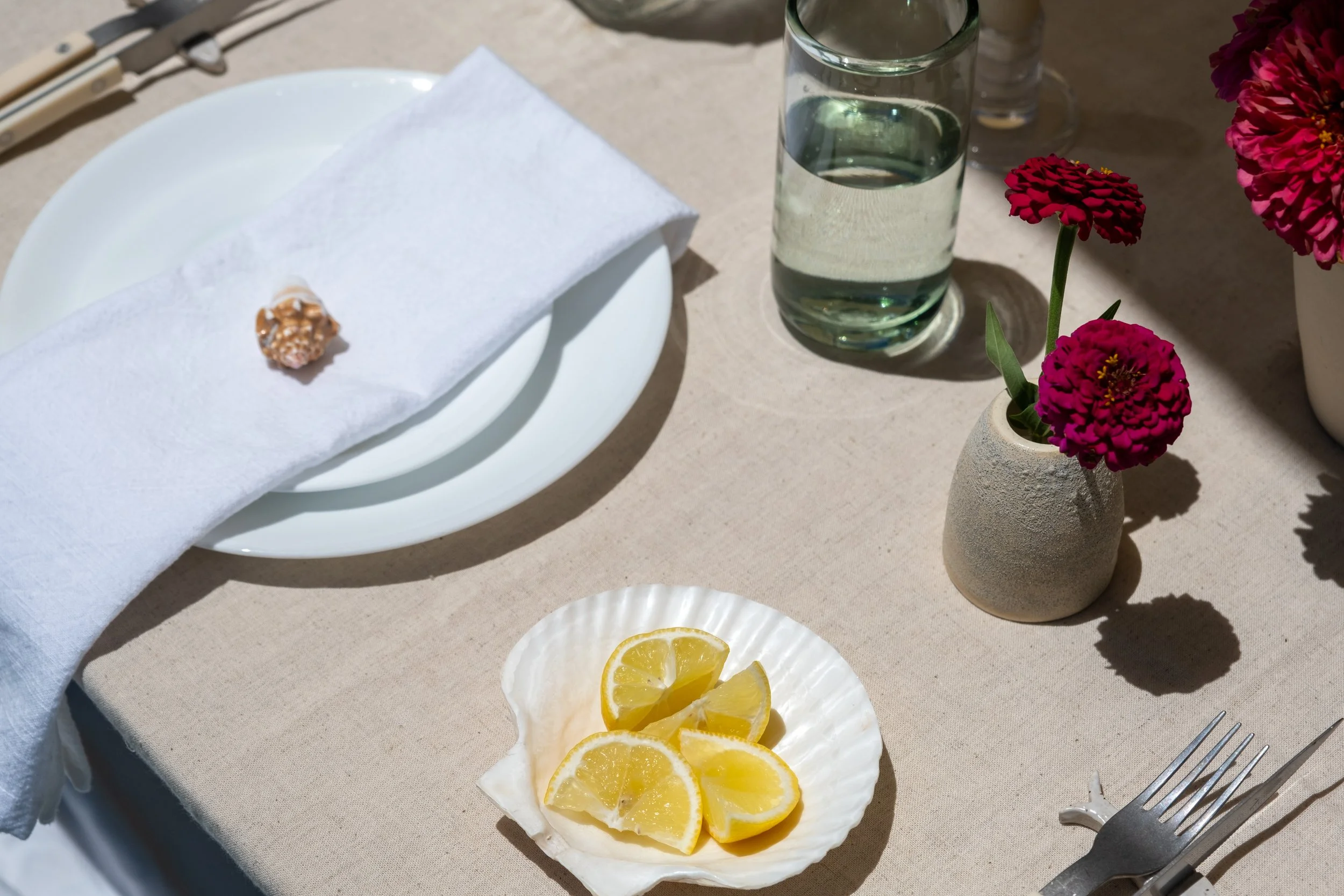 A table setting with a white plate, a napkin with a piece of bread, a small bowl of lemon wedges, a glass of water, and a small vase with pink and red flowers.